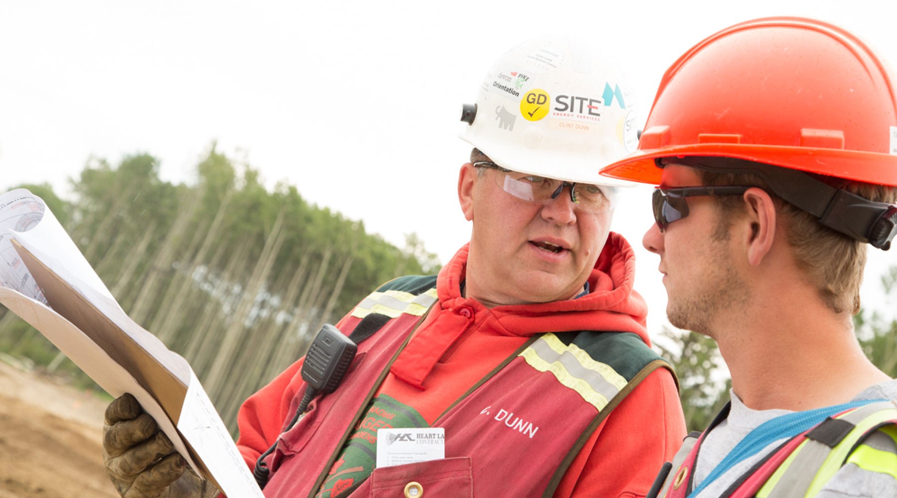 Image of two workers wearing PPE and hardhats, holding papers and conversing Image of two workers wearing PPE and hardhats, holding papers and conversing