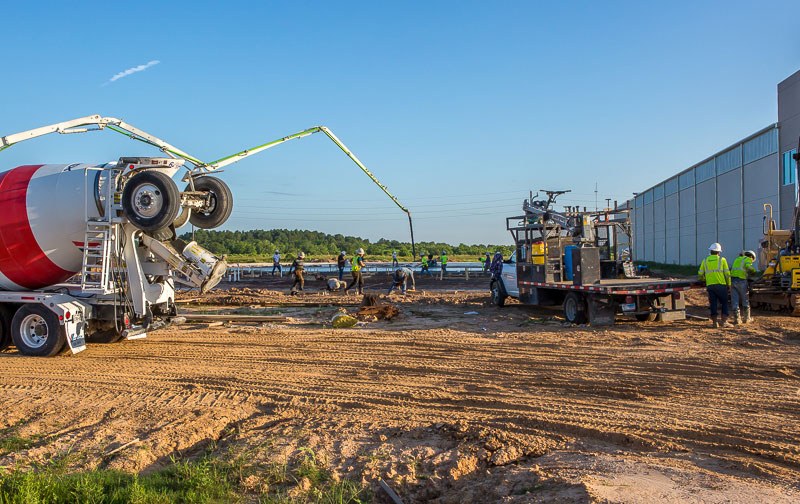 Image of a cement truck and flatbed truck on a construction site with several workers in the background